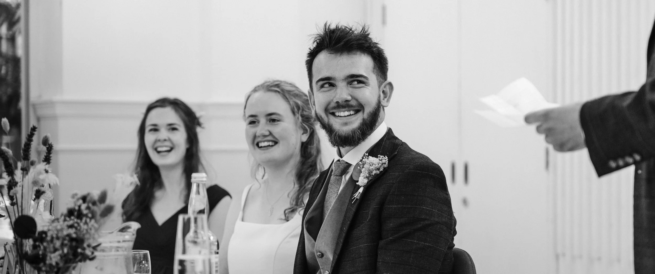 Black and white photo of three smiling young adults, two women and one man, sitting at a table during a formal event, with a person in a suit reading from a paper on the right.