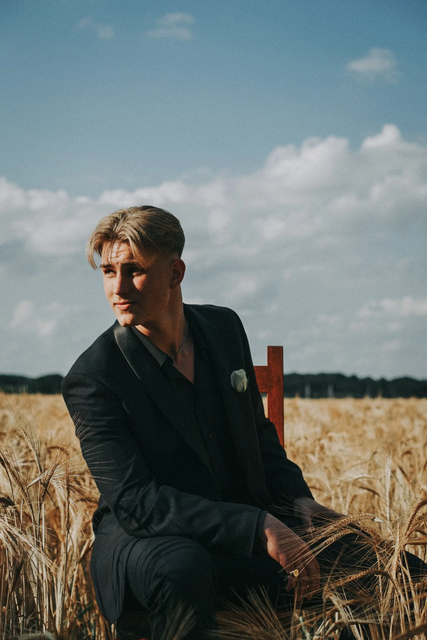 A young man in a black suit is sitting on a red chair in a wheat field under a partly cloudy sky.