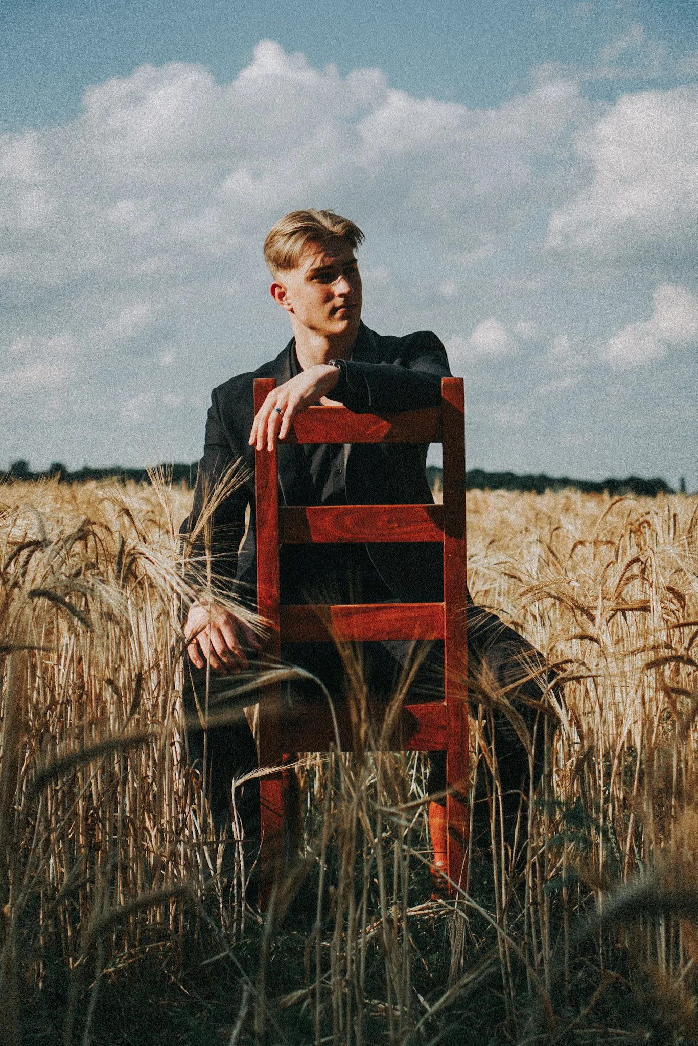 A young man in a black suit sitting on a wooden chair amid a wheat field under a partly cloudy sky.