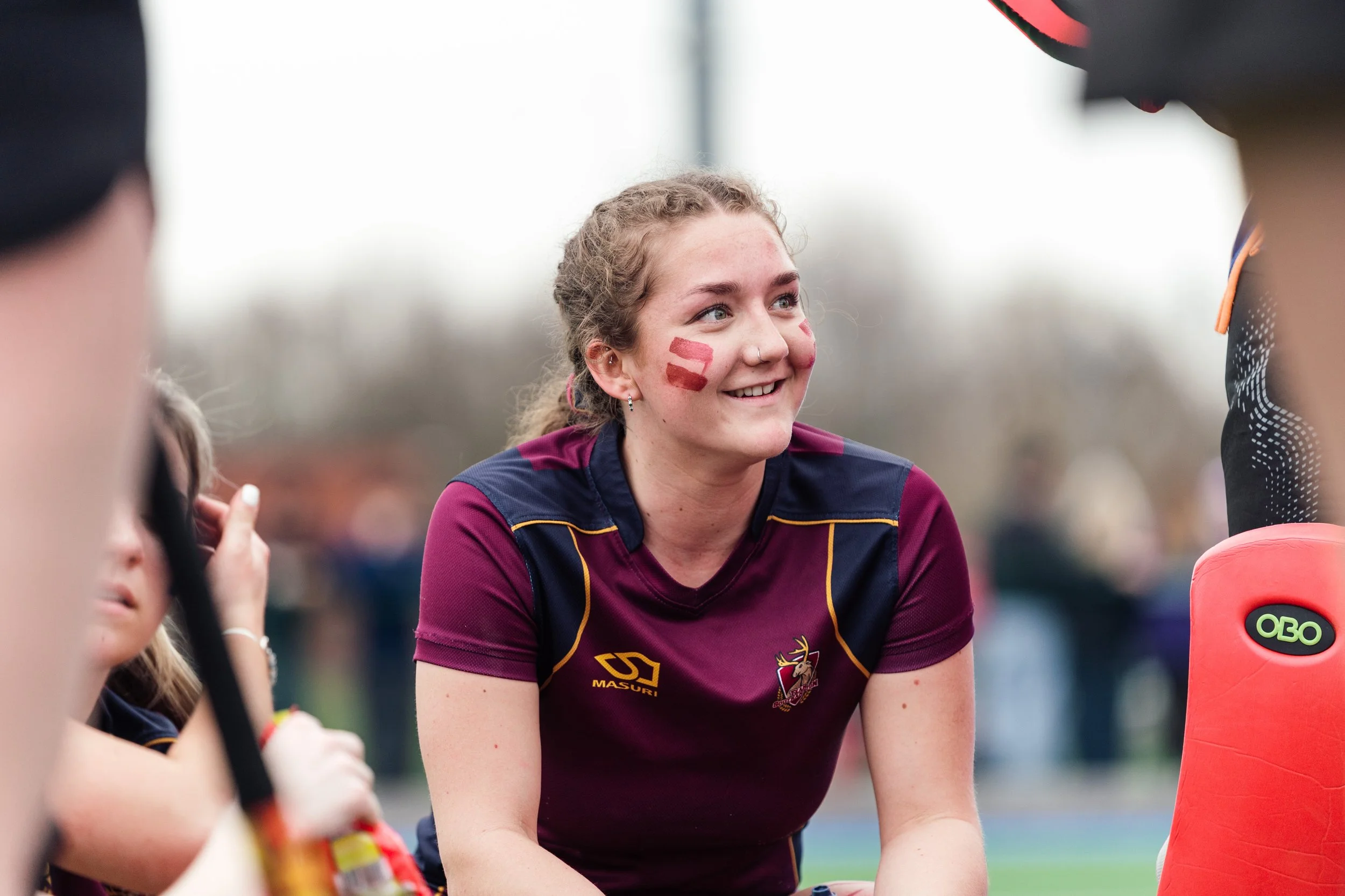 A young woman with curly hair and face paint on her cheek smiling while sitting at a sports event, wearing a maroon and navy sports jersey, surrounded by others.