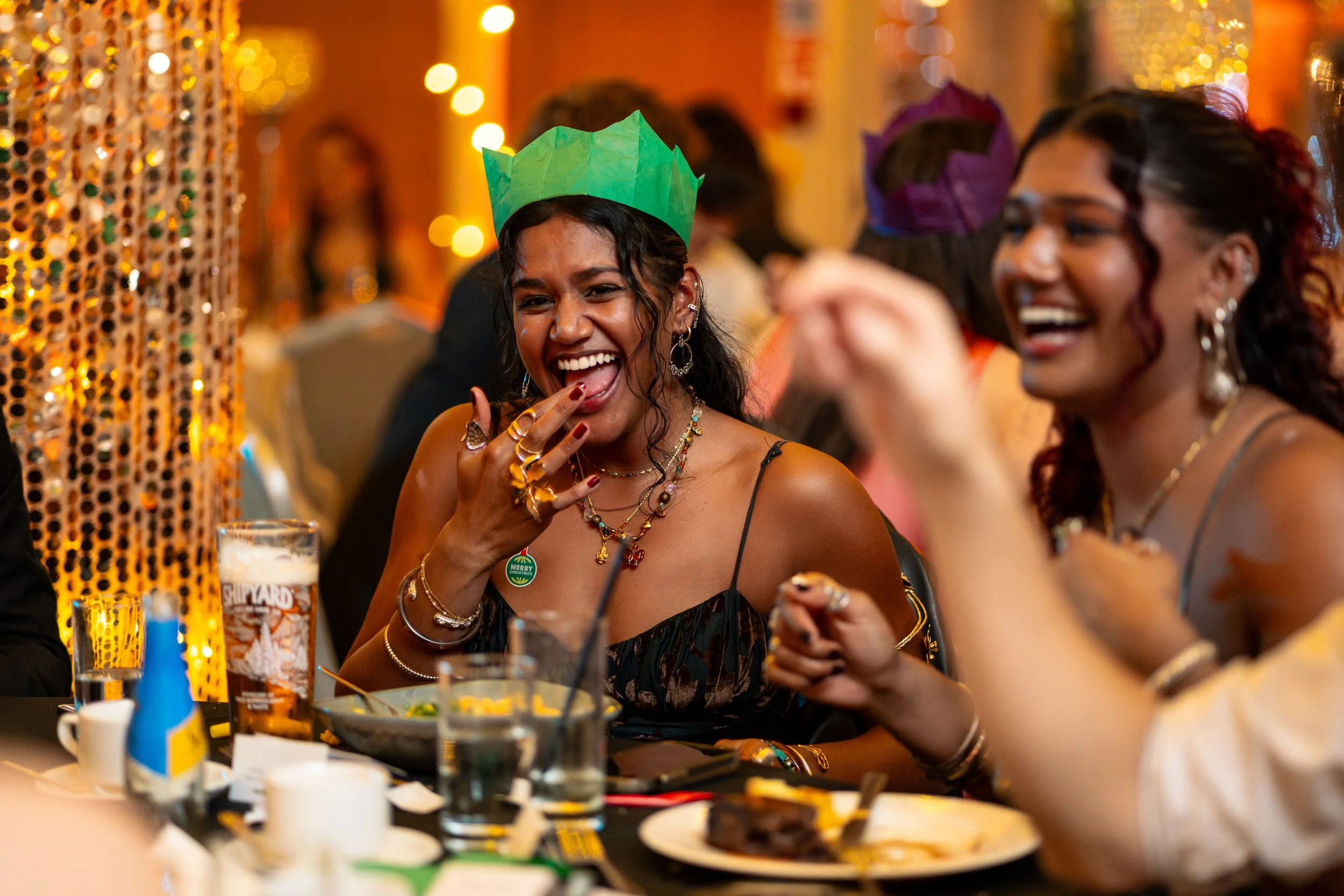 Women celebrating, wearing colorful party hats, smiling and laughing at a lively party or gathering with warm lighting and festive decorations.