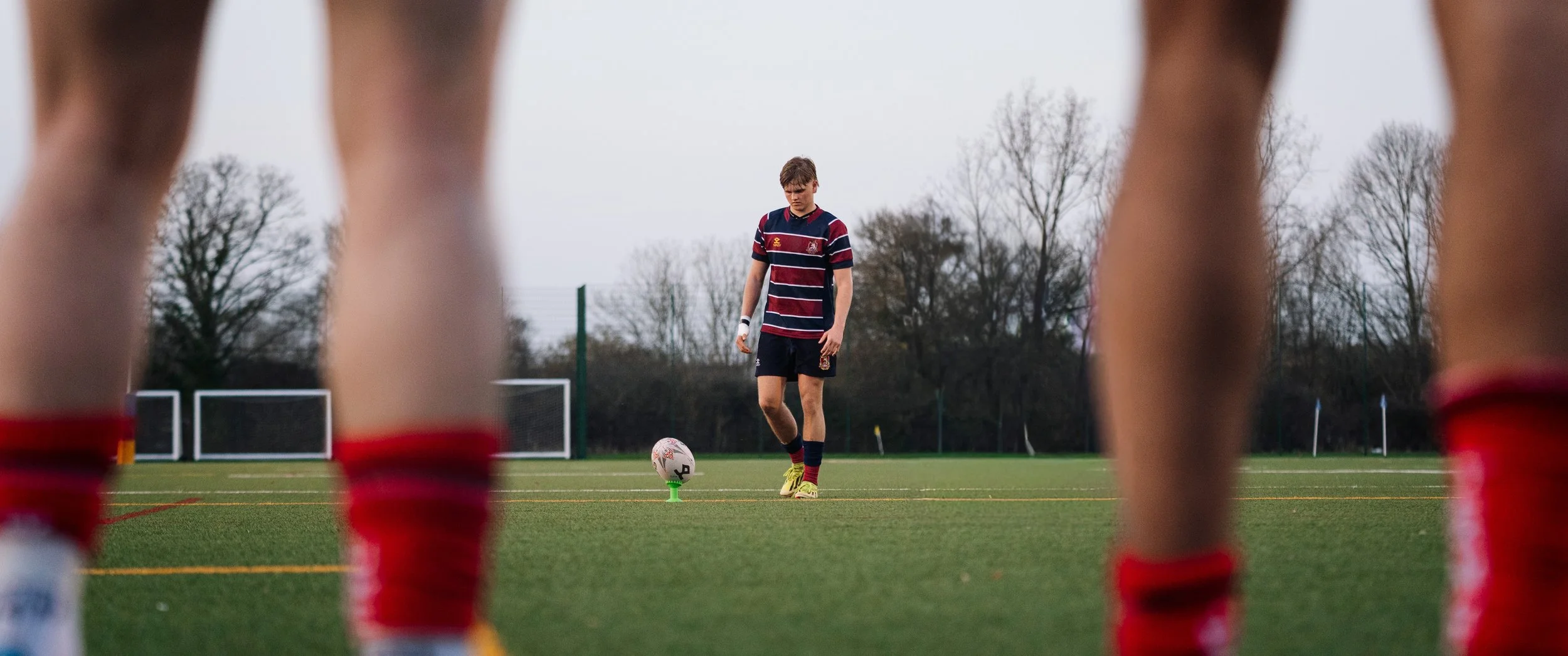 A young male soccer player standing on a field preparing to kick a ball, viewed through the legs of other players wearing red socks.