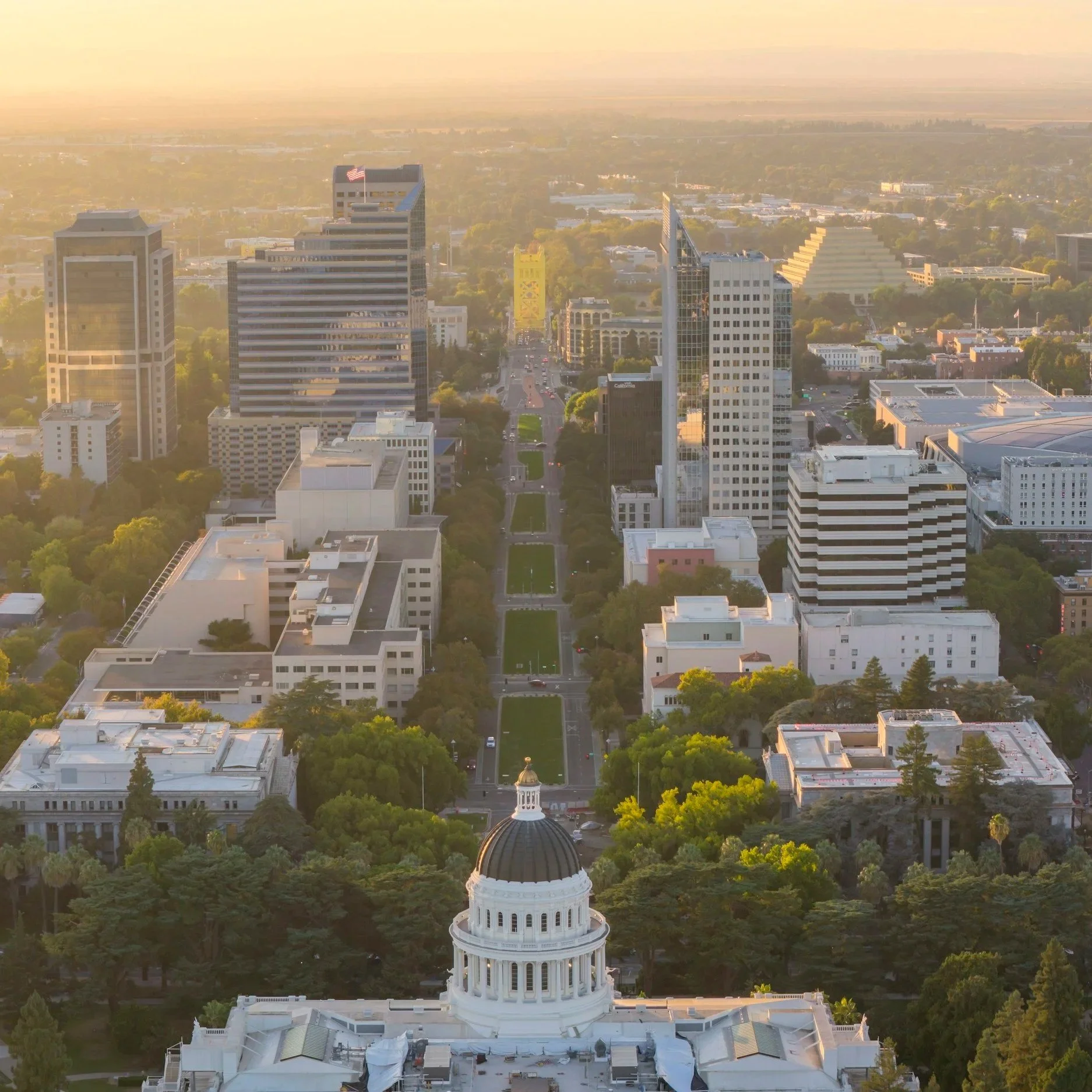 Aerial view of a city skyline at sunset with tall buildings, a historic domed capitol building in the foreground, and lush greenery surrounding the area.