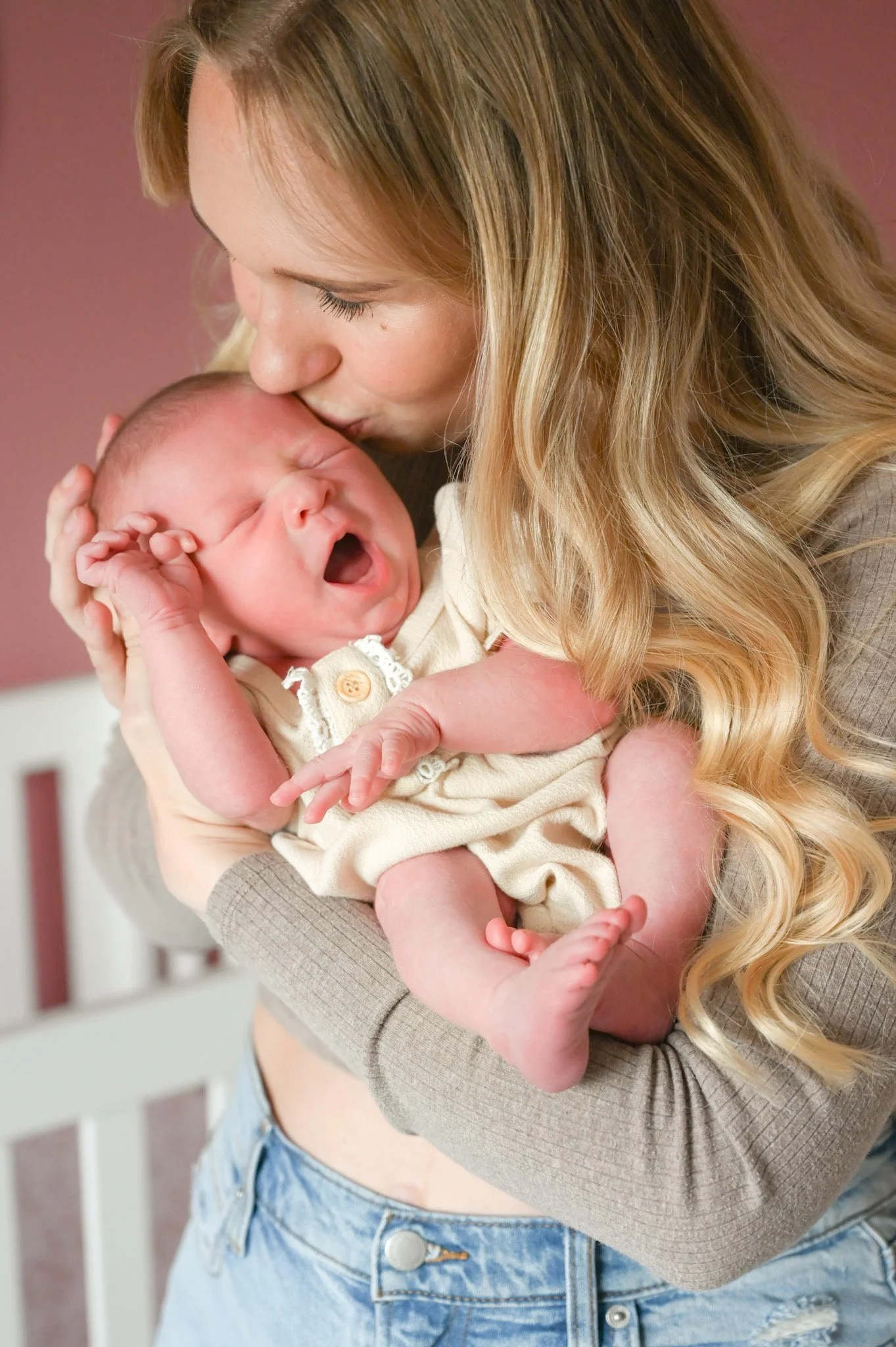 A woman with long blonde hair holding a yawning baby with light skin and a beige outfit.