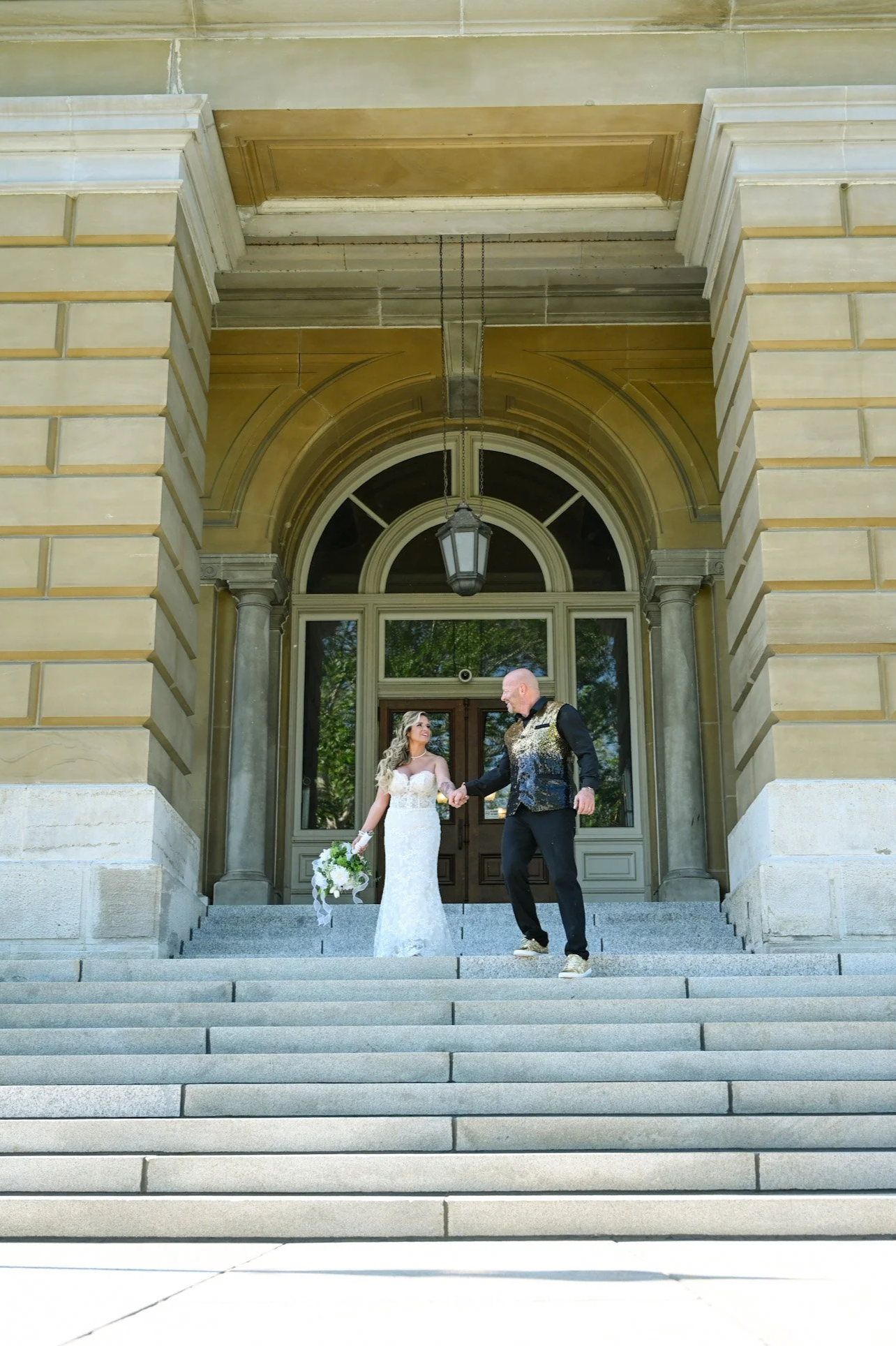 A bride and groom walking down the steps of a large, historic building with columns and an arched entrance, holding hands and smiling at each other. The bride is holding a bouquet of flowers.