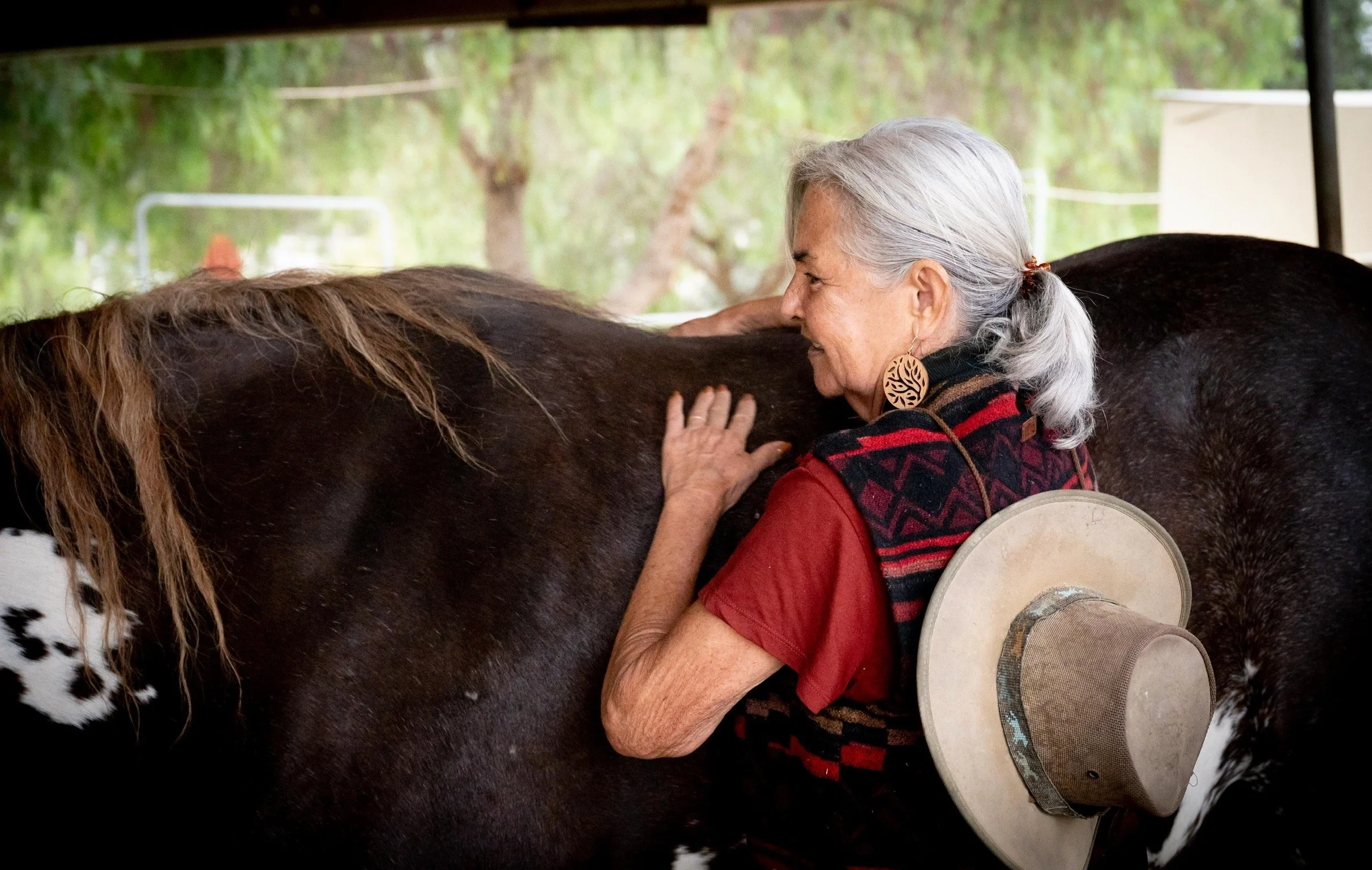 An elderly woman with silver hair, wearing a red shirt, a patterned vest, and large earrings, is hugging a large brown and black horse. She has a wide-brimmed hat hanging from her arm, and is standing in a green outdoor setting with trees in the background.