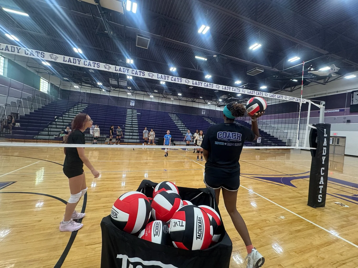 A coach holding a volleyball while athletes prepare for a drill.