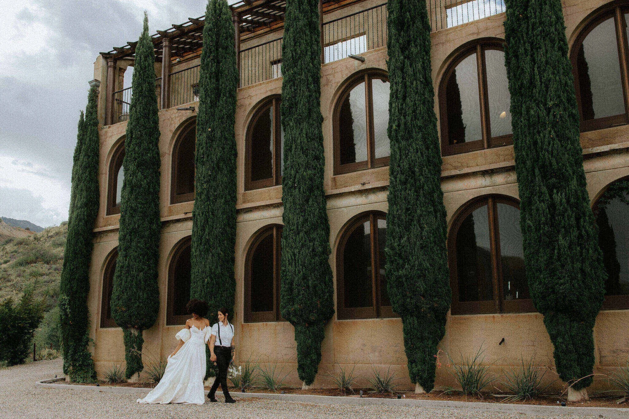 2 brides walking in front of a tuscany inspired building with cypress trees