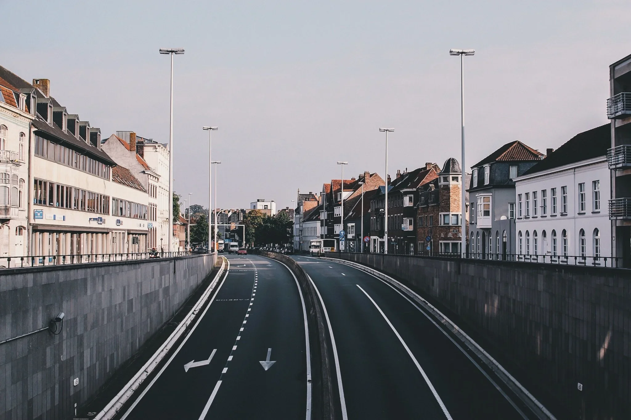 Empty urban street with buildings on both sides, streetlights, and tram tracks, under a cloudy sky.