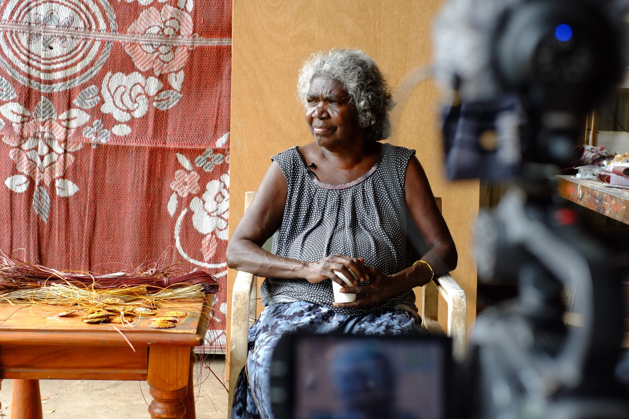An elderly woman with gray curly hair, wearing a black and white polka dot sleeveless top and a patterned skirt, sitting in a chair holding a small cup. To her left, a table with woven jewelry or craft items. The background features a red floral quilting or tapestry and a plain wall.