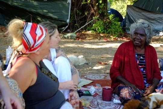 Two women sitting outdoors with an Aboriginal Elder on a blanket, engaged in conversation, with trees and tents in the background.