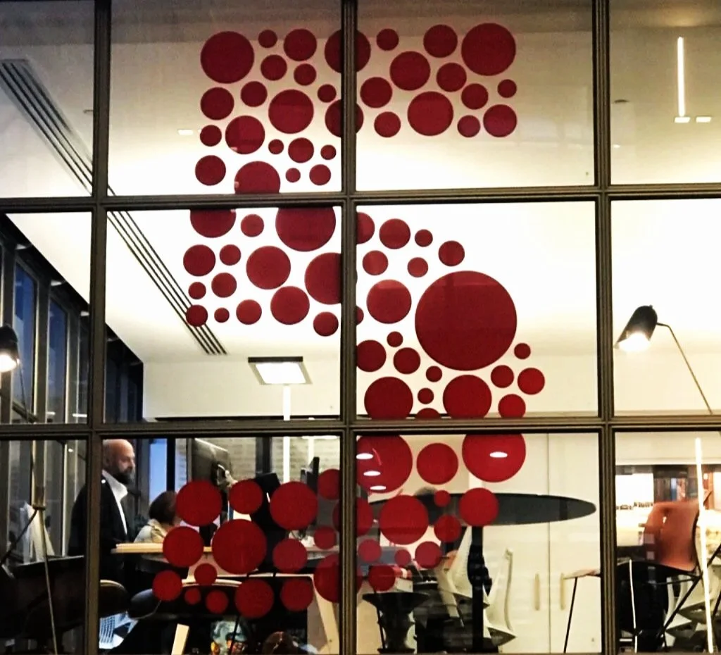 A glass window with red circular decals forming a pattern inside a modern cafe or office, with people working and chatting inside.