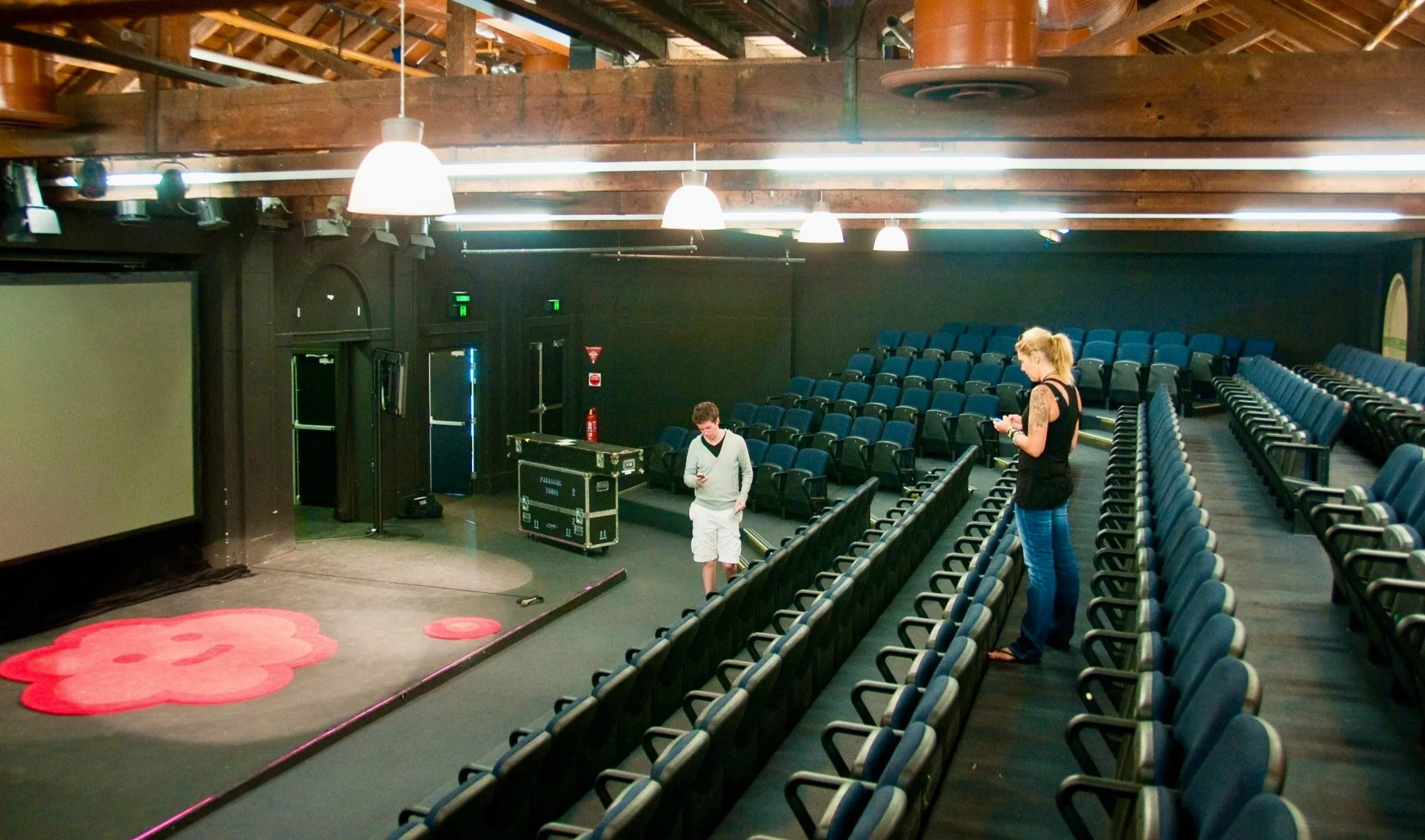 Empty theater with rows of blue seats, a small stage with a pink flower design, two people checking their phones, and a large screen on the left.