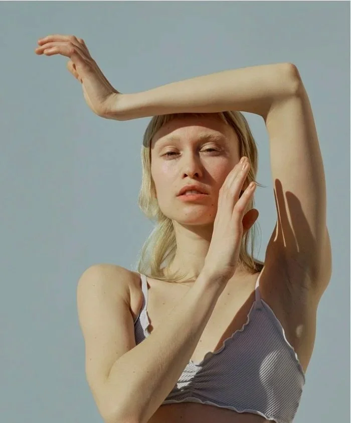 A woman with light blonde hair is outdoors against a pale blue sky, posing with one arm raised above her head and the other hand touching her face. She is wearing a sleeveless, striped top.