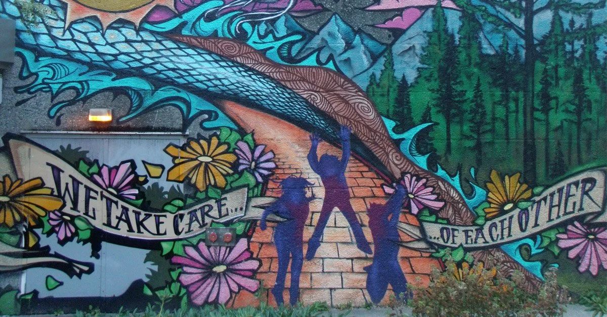 Colorful mural depicting three silhouettes of people holding hands underneath a large blue fishing rod, surrounded by flowers, mountains, and trees, with a banner that reads "We take care... of each other."