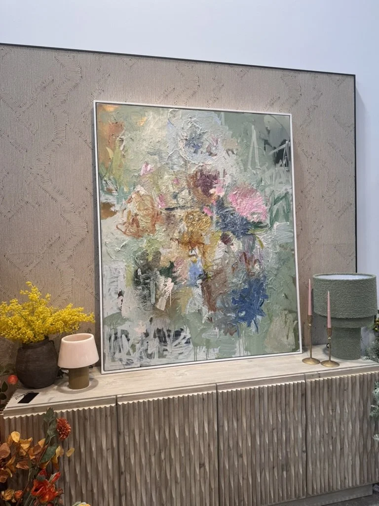 Decorative interior with a large abstract floral painting on a textured beige wall, a table with yellow flowers, a pink and beige table lamp, green textured lampshade, pink candlesticks, and orange flowers.