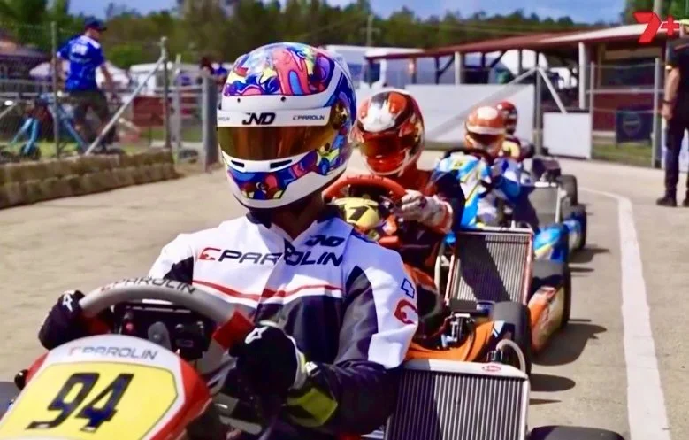Three go-kart racers in line at the starting grid, wearing helmets and racing suits, with a racing track and spectators in the background.