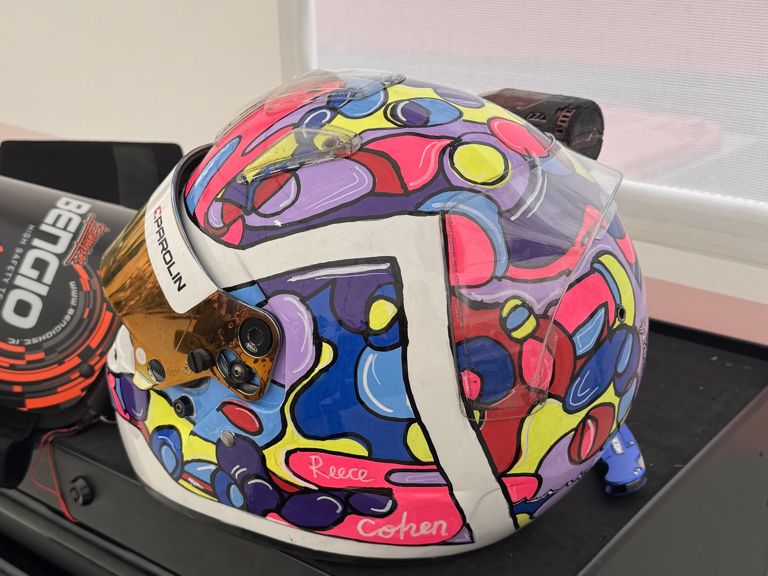 A motorcycle helmet with a colorful, abstract, graffiti-style design in pink, yellow, blue, purple, and black, placed on a desk next to a pair of headphones.