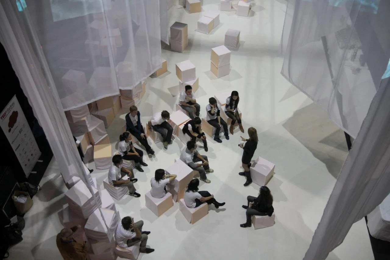Group of people attending a presentation or workshop in a modern, minimalist space with sheer curtains and seating cubes.