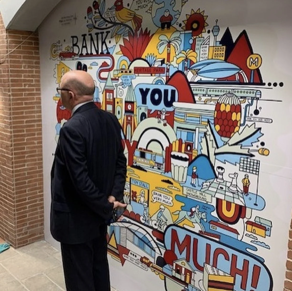 A man in a suit with glasses stands with his back to the camera, inside a building with brick walls, in front of a colorful mural depicting a cityscape with various landmarks, illustrations, and the words 'YOU' and 'MUCH' prominently displayed.