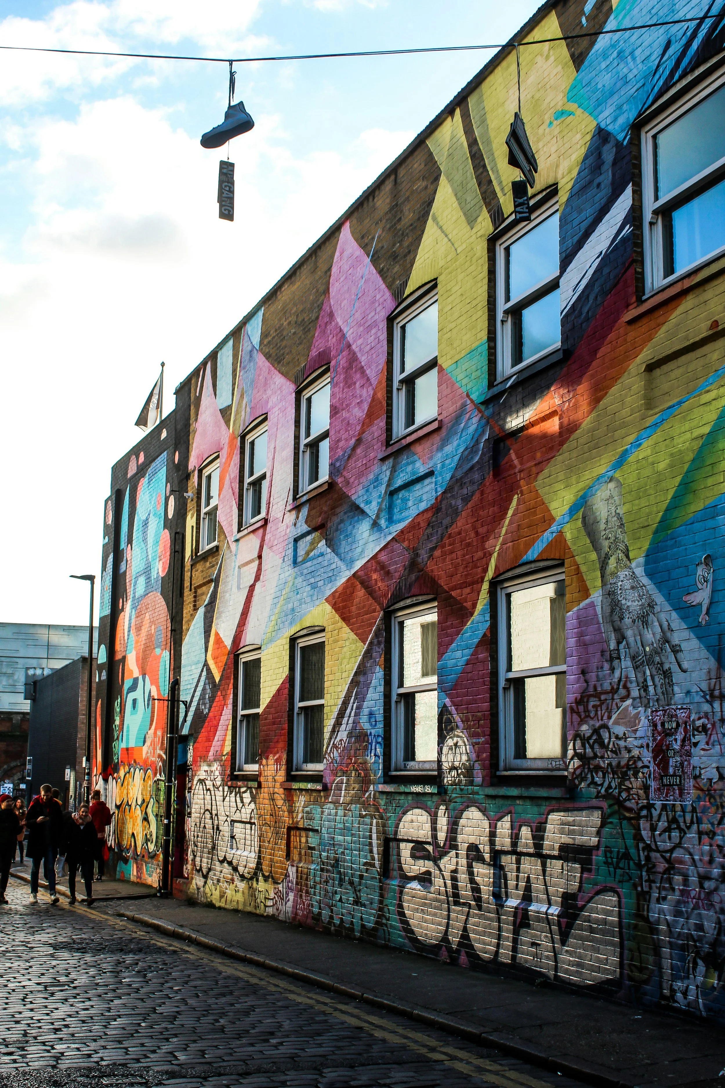Street view of a colorful mural painted on a brick building with several windows; graffiti and a small group of people walking on the cobblestone sidewalk.