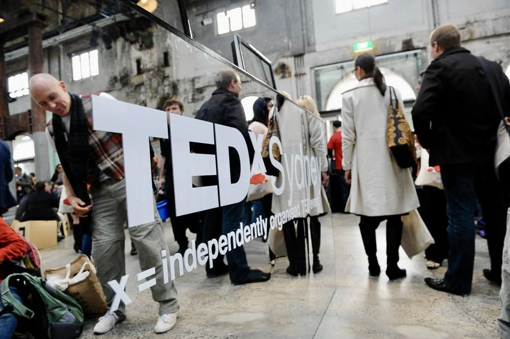 People gathered at a TEDx event, standing behind a reflective glass wall with a large TEDx sign on it, inside an industrial-style venue with exposed brick and cement walls.