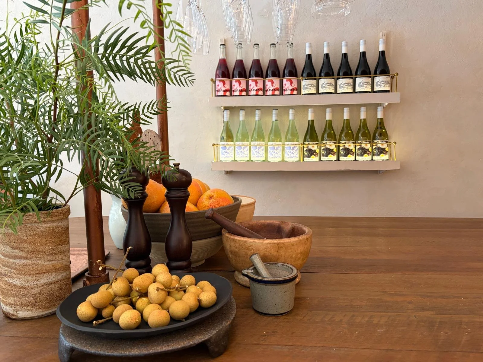 A wooden table with a potted plant, a plate of longan fruit, a bowl of oranges, and various kitchen items. A wall shelf holds bottles of wine, and hanging glassware is above the shelf.