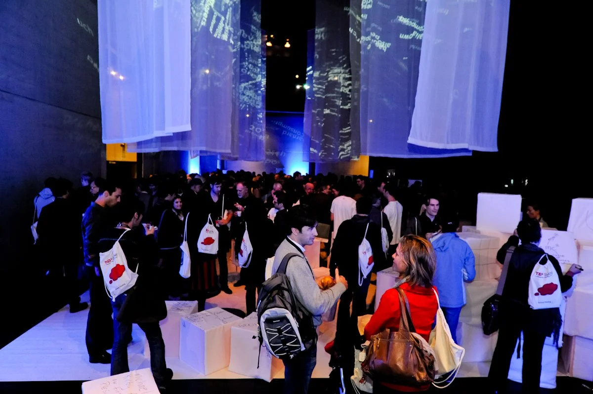 People gathered at TEDxSydney, many carrying tote bags, in a dimly lit room with large, semi-transparent hanging fabric panels and projected digital text on the ceiling.