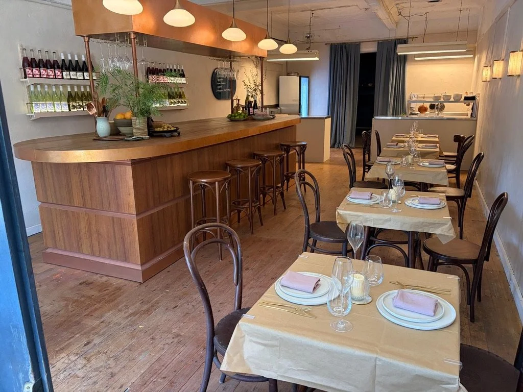 Interior of a restaurant with tables set for dining, a wooden bar with stools, and wine bottles displayed on shelves.