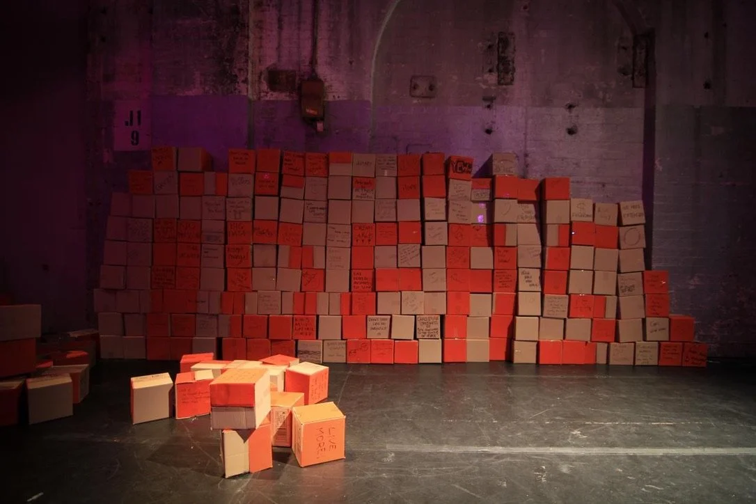 A wall at TEDxSydney made of stacked cardboard boxes with handwritten messages, in a dimly lit room with purple lighting and a brick wall background.