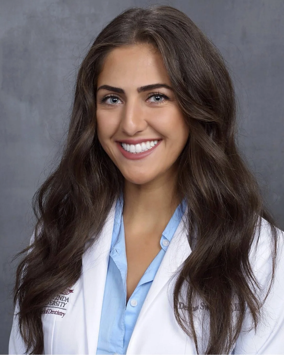 A smiling woman with long brown hair and blue eyes wearing a white coat over a light blue shirt against a gray background.