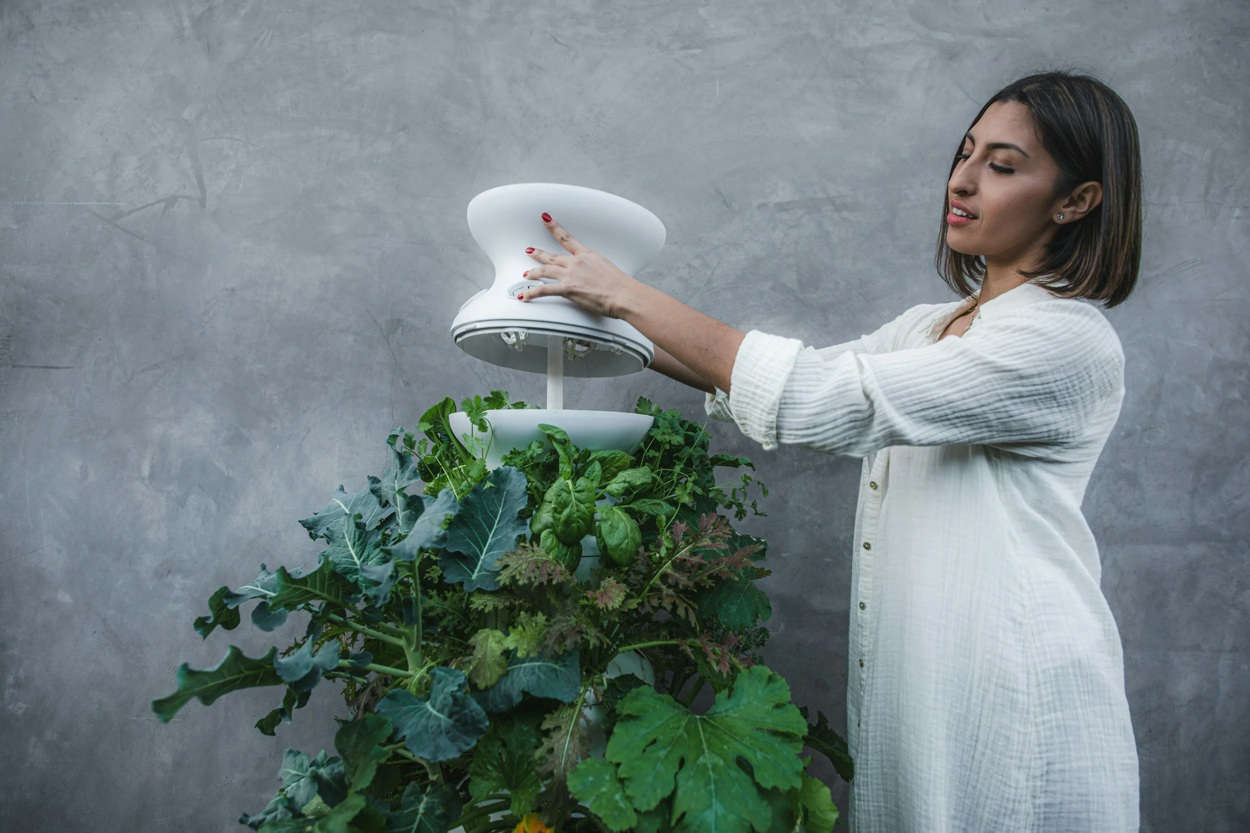 A woman in a white shirt harvests vegetables from a vertical garden with a white, modern gardening device against a gray background.