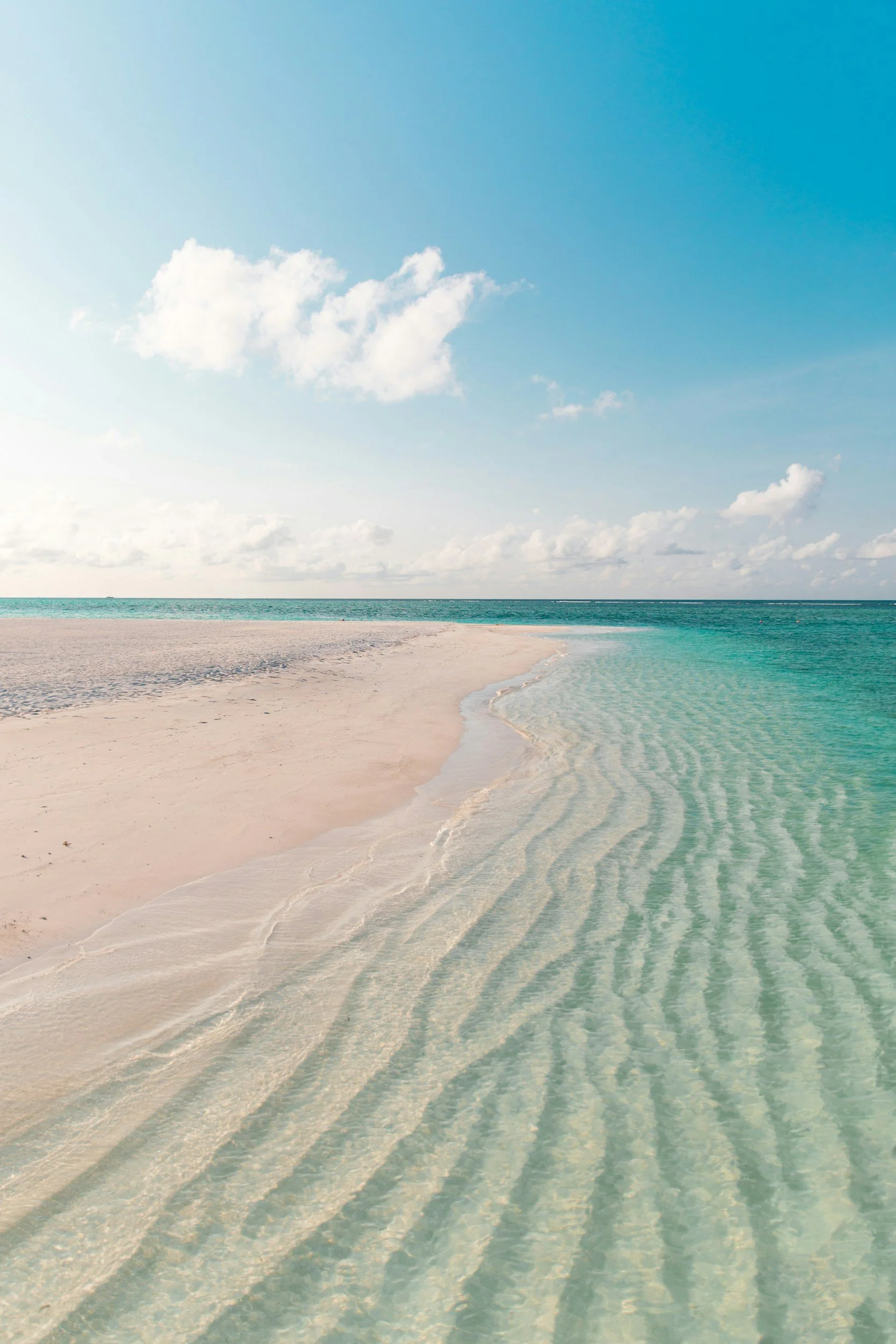 A serene beach scene with clear turquoise water gently lapping against white sandy shore, under a bright blue sky with scattered white clouds.