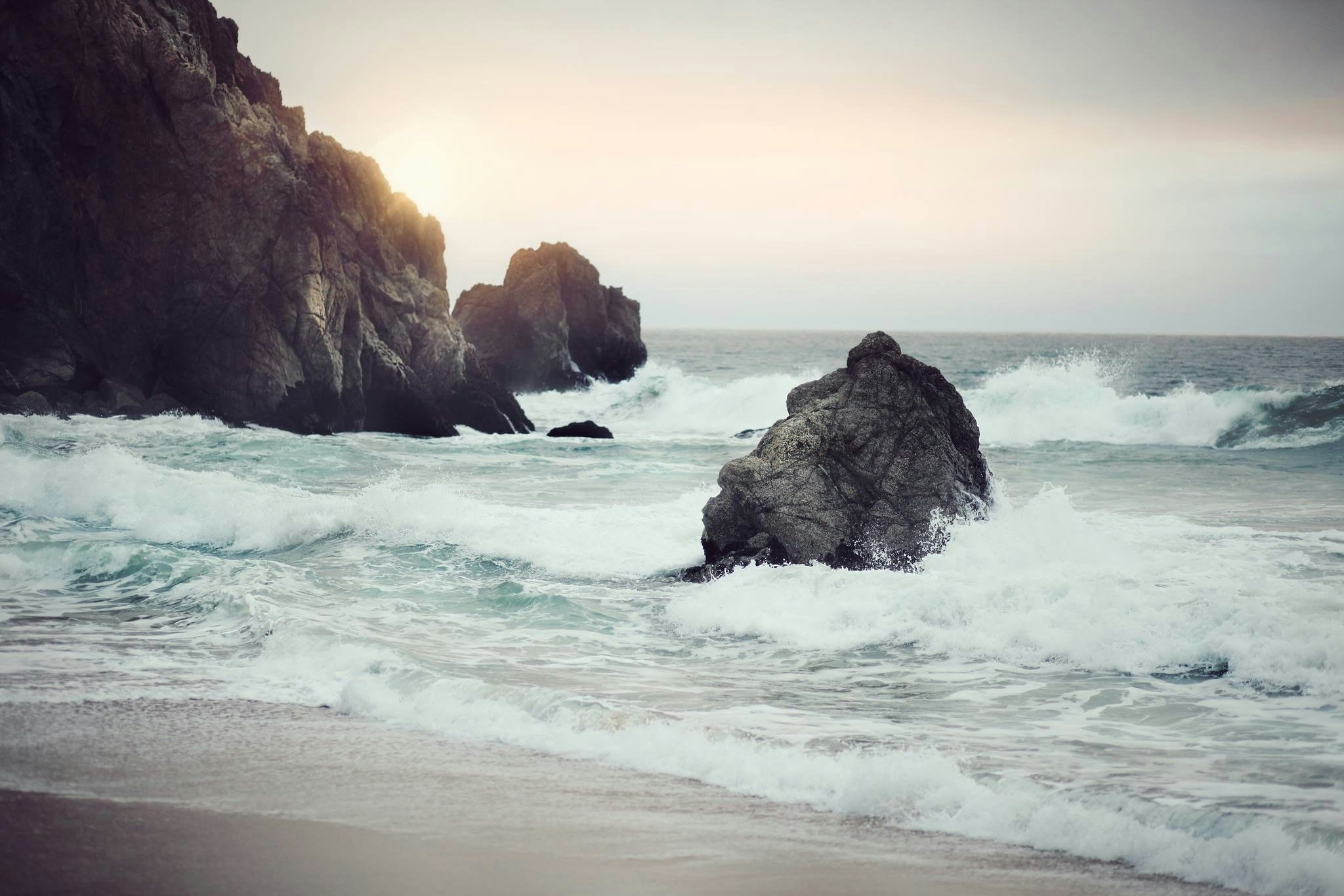 Waves crashing against rocks on a beach at sunset with cliffs and cloudy sky in the background.