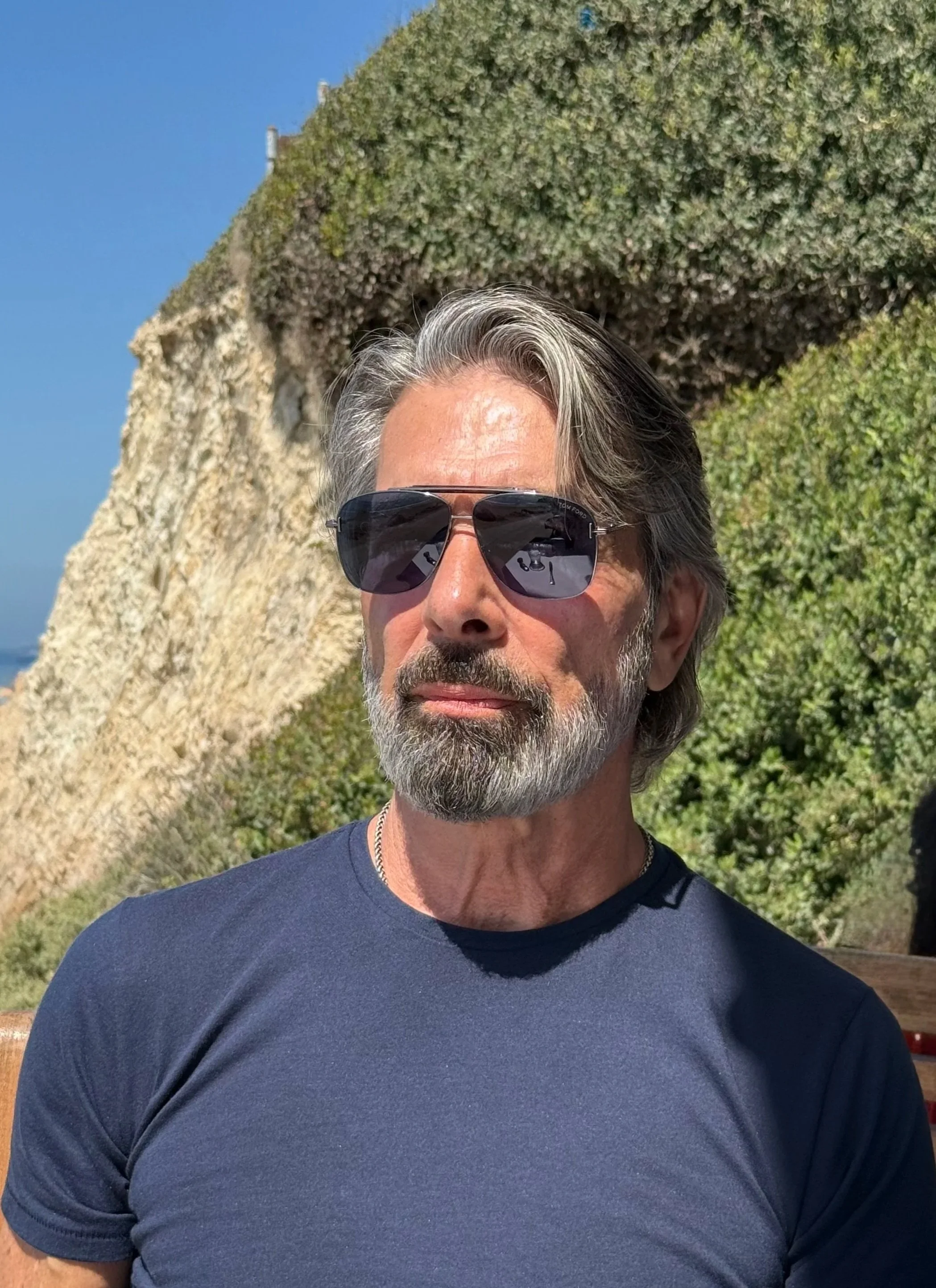 A man with gray hair and a beard wearing sunglasses and a navy blue t-shirt, sitting outdoors with a rocky cliff and green bushes in the background.