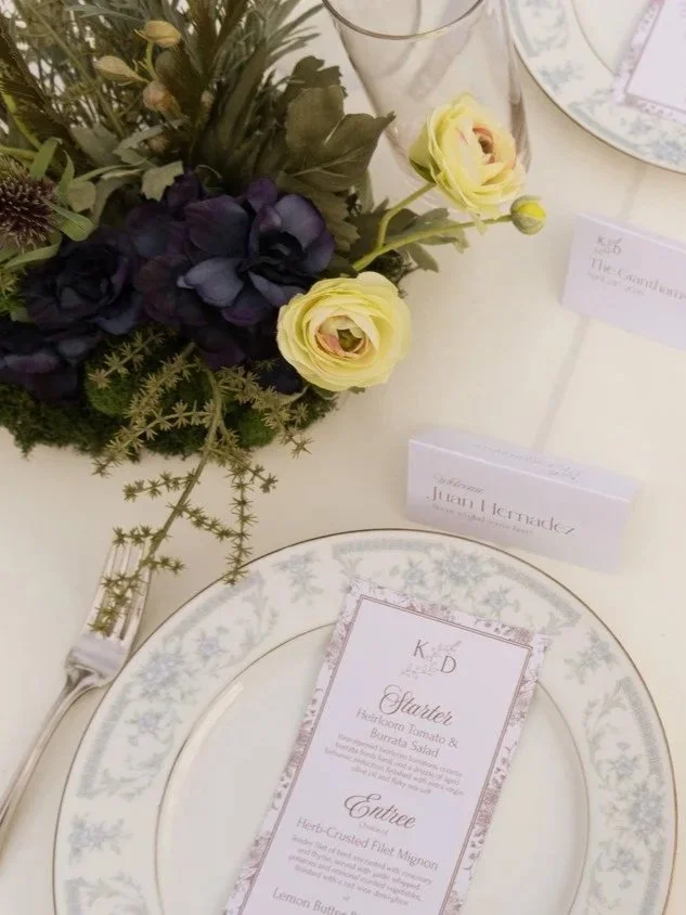 Elegant wedding reception table setting with a purple and yellow floral centerpiece and a custom menu card featuring burrata salad and herb-crusted filet mignon.