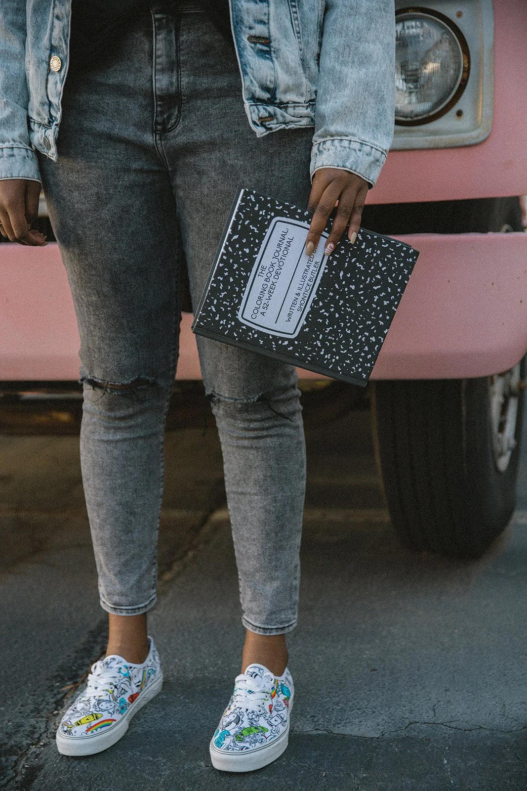 A person wearing a denim jacket and jeans, holding a black and white composition notebook, standing in front of a pink vehicle with a displayed washer or dryer.