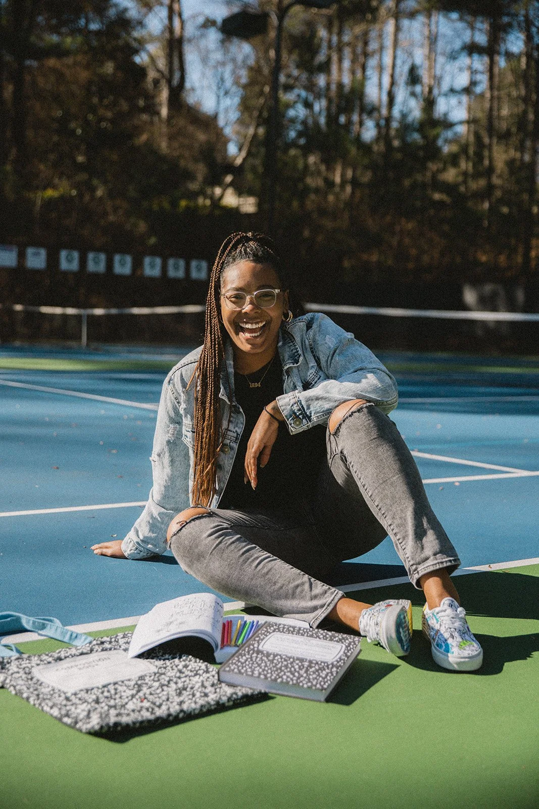 A young woman sitting on the ground outdoors on a tennis or basketball court, smiling and laughing. She has long braids, glasses, a denim jacket, ripped jeans, and colorful sneakers. There are notebooks, colored markers, and a bag on the ground in front of her.
