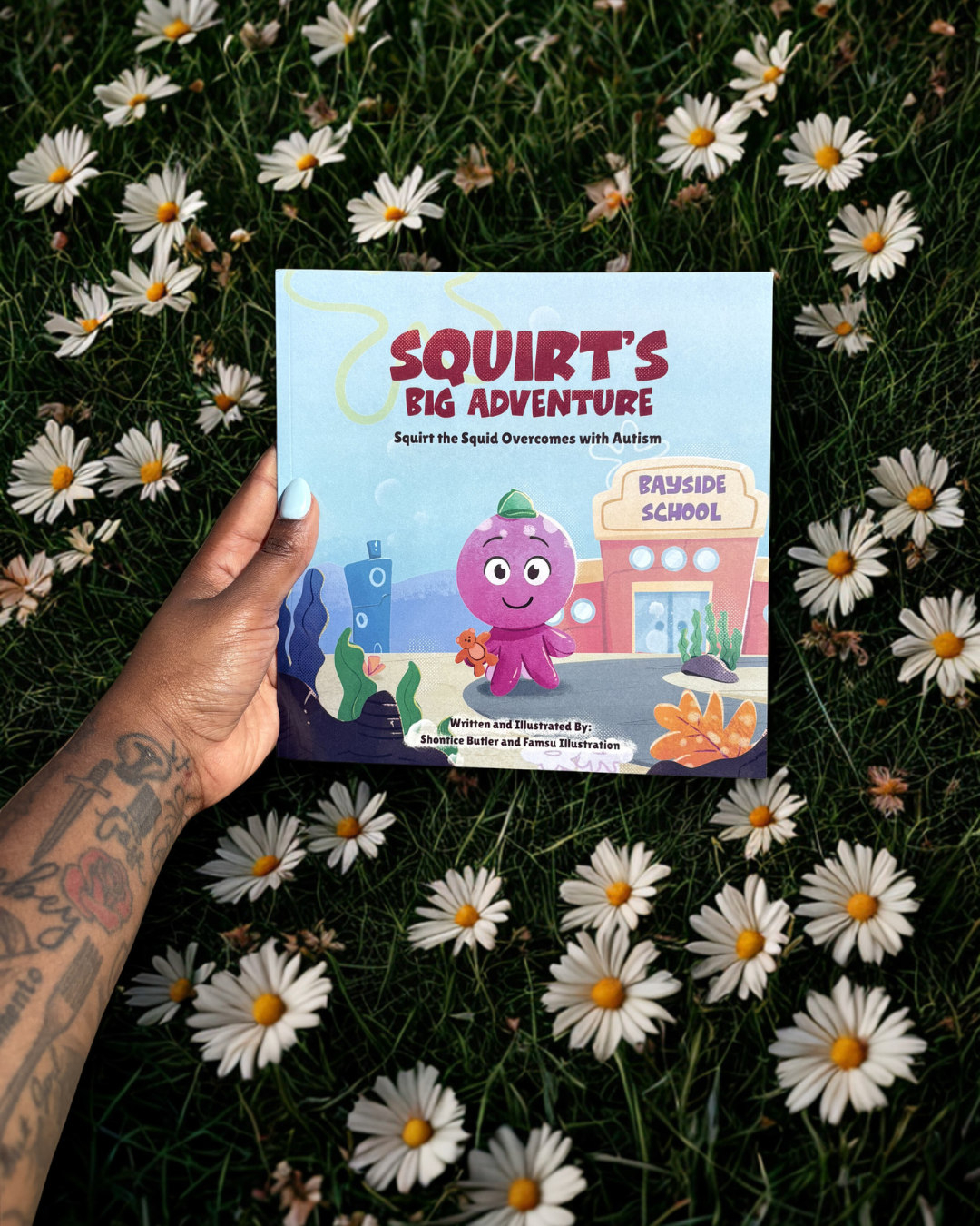 A children's book titled 'Squirt's Big Adventure' held over a field of white daisies with yellow centers. The book's cover features a purple squid cartoon squirt with a green hat, local scenery, and a sign that reads 'Bayside School.'
