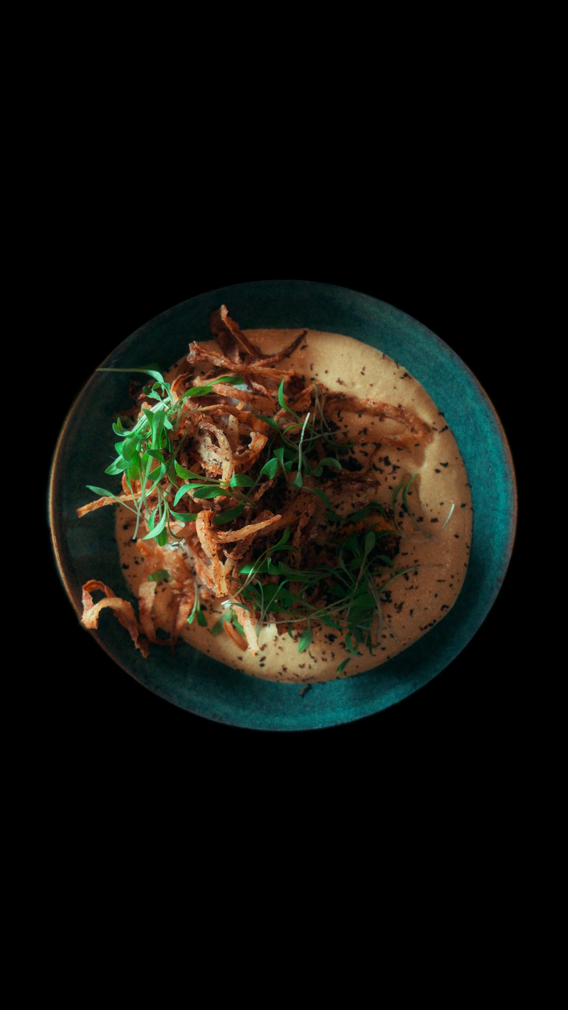 Prato azulado com bacalhau, creme de cebola com cebolinha, cebolas fritas crocantes e ervas verdes.