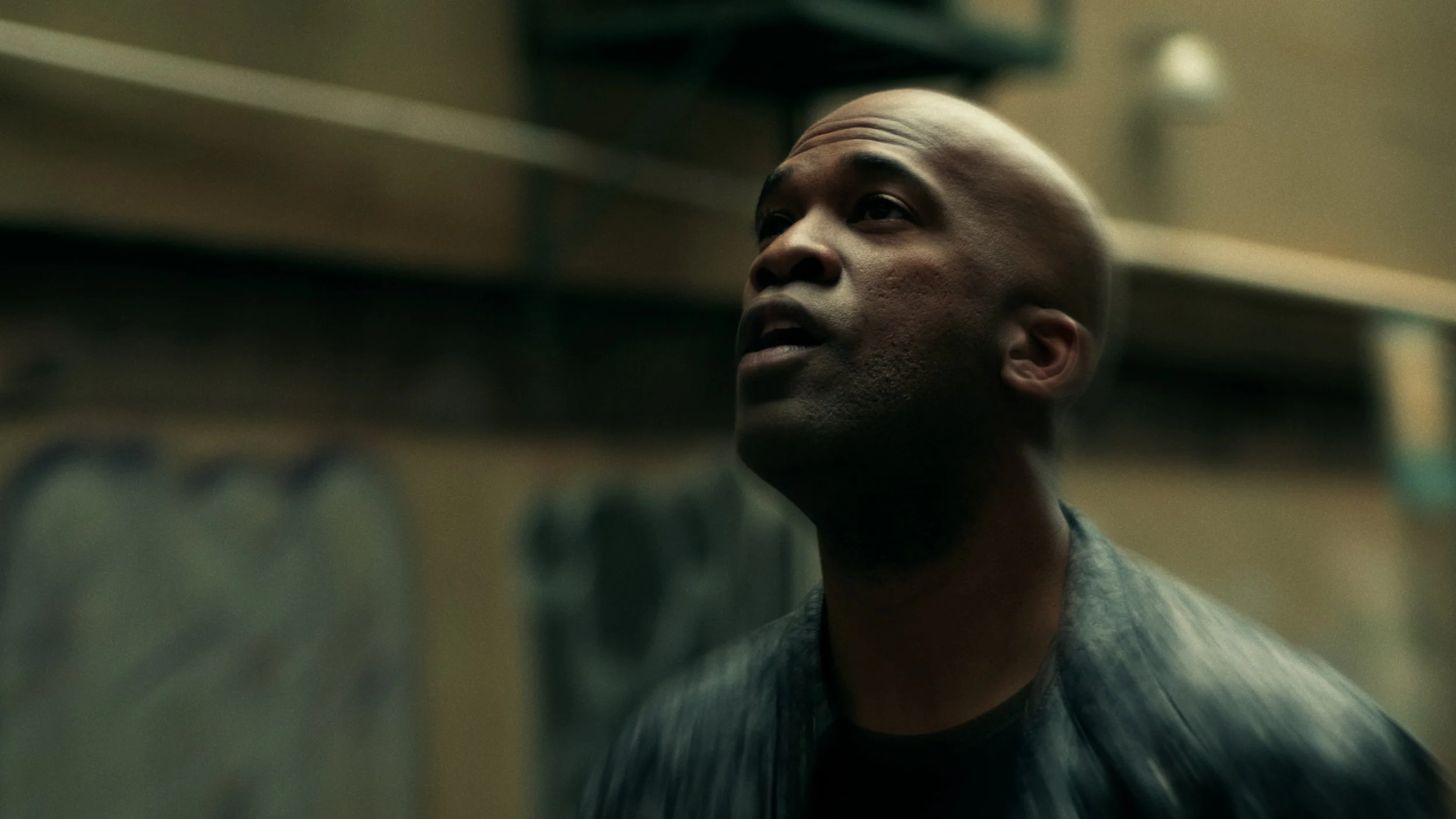 Close-up de um homem negro com cabeça raspada, olhando para cima, no interior de um local com prateleiras e garrafas ao fundo.