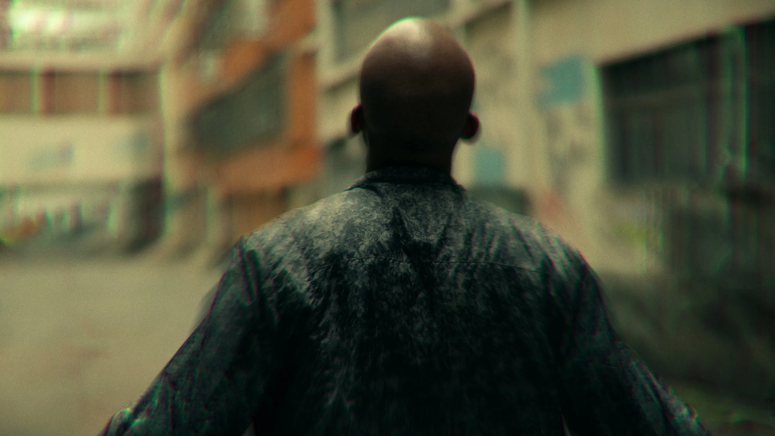 Pessoa vista de costas, com cabelo raspado, usando uma jaqueta escura, caminhando por uma rua urbana, com prédios ao redor.