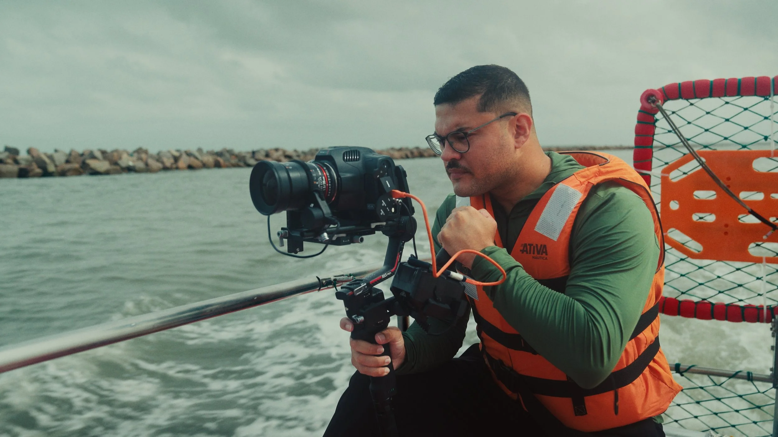 Homem operando câmera de vídeo em embarcação no mar, usando colete salva-vidas laranja.