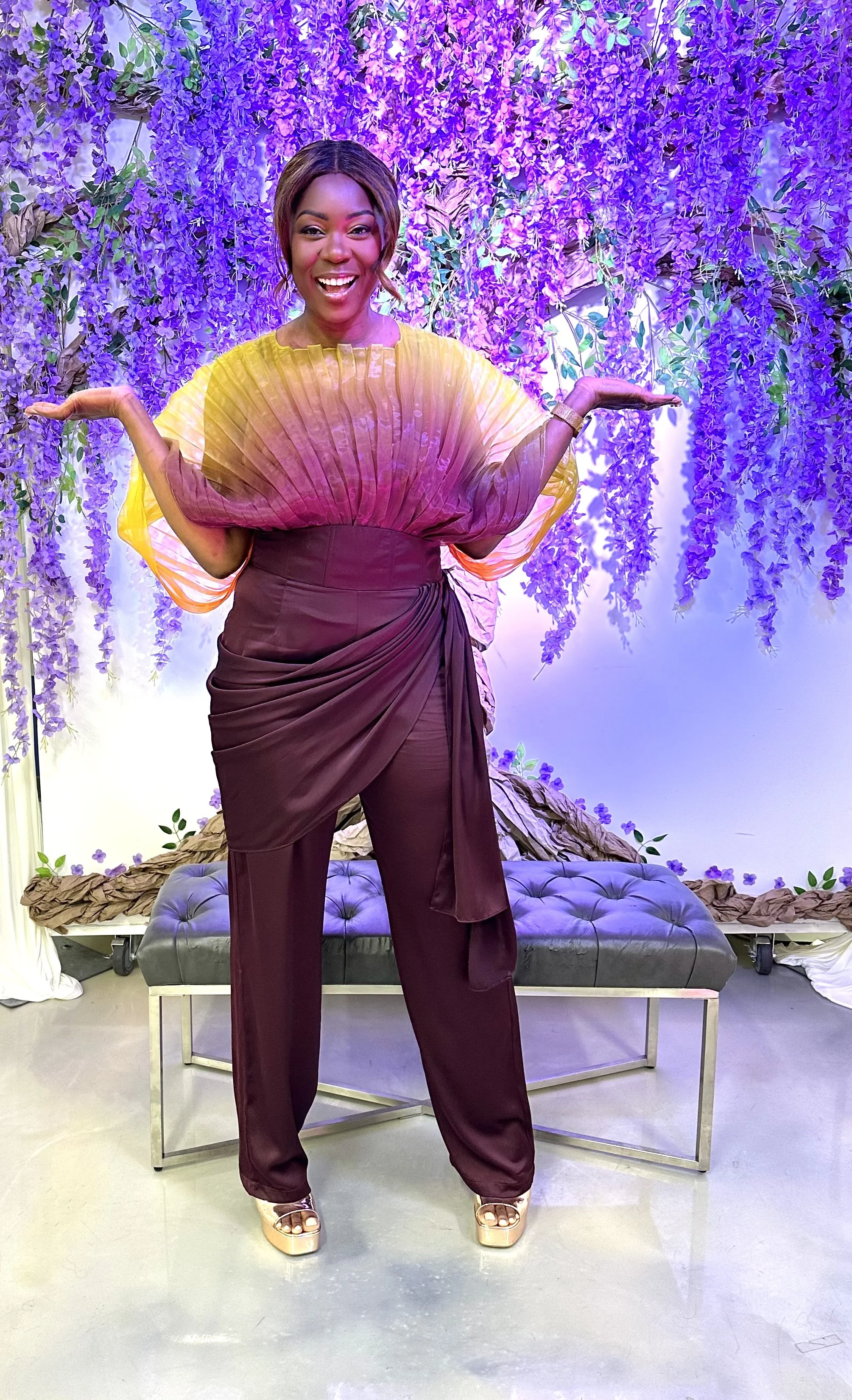 A woman with short hair smiling and posing with her arms outstretched in front of a purple floral backdrop with hanging flowers, standing on a polished floor in front of a bench.