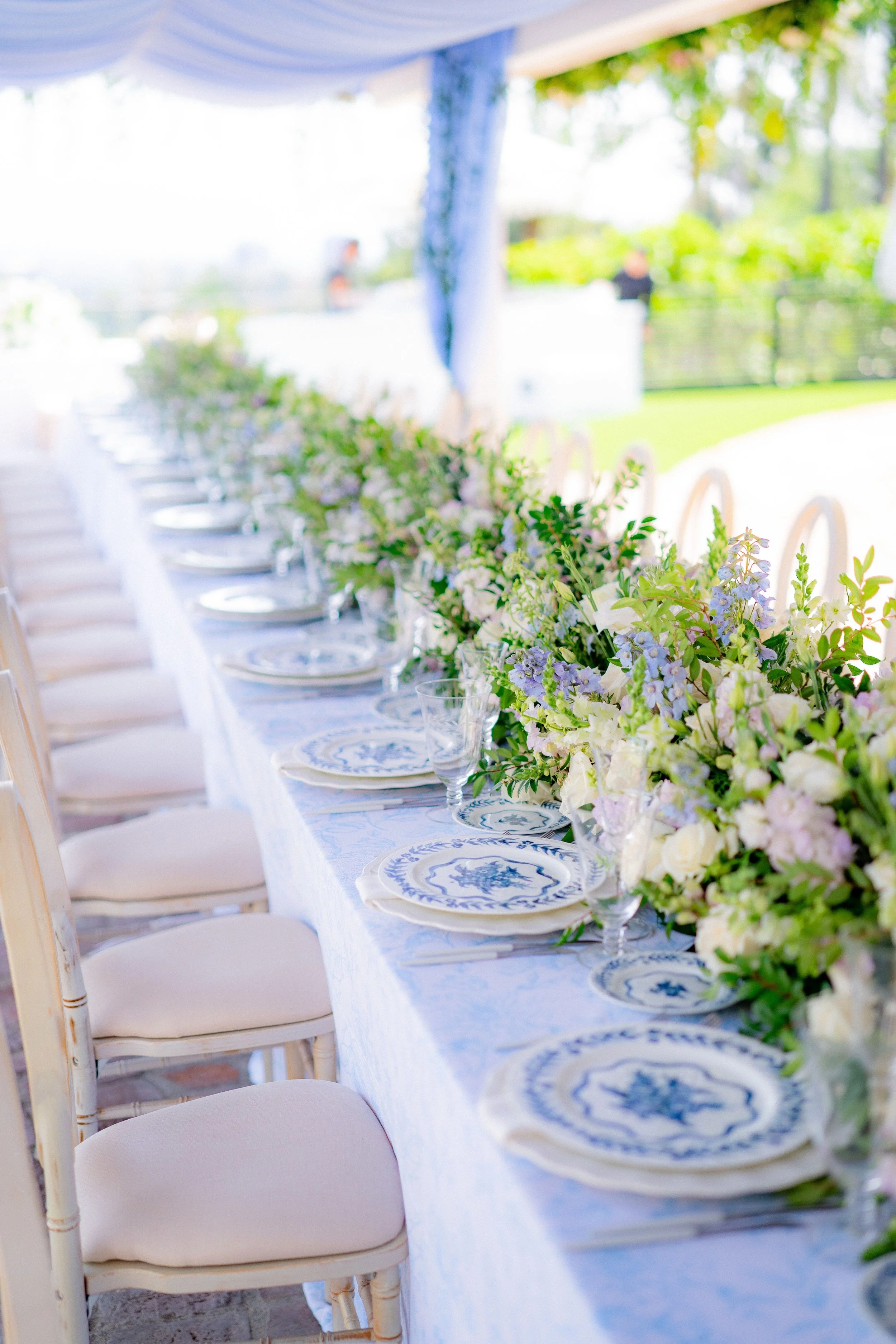 Long banquet table set for an outdoor event with floral centerpieces, blue and white patterned plates, clear glassware, and white cushioned chairs, under a white tent with greenery in the background.