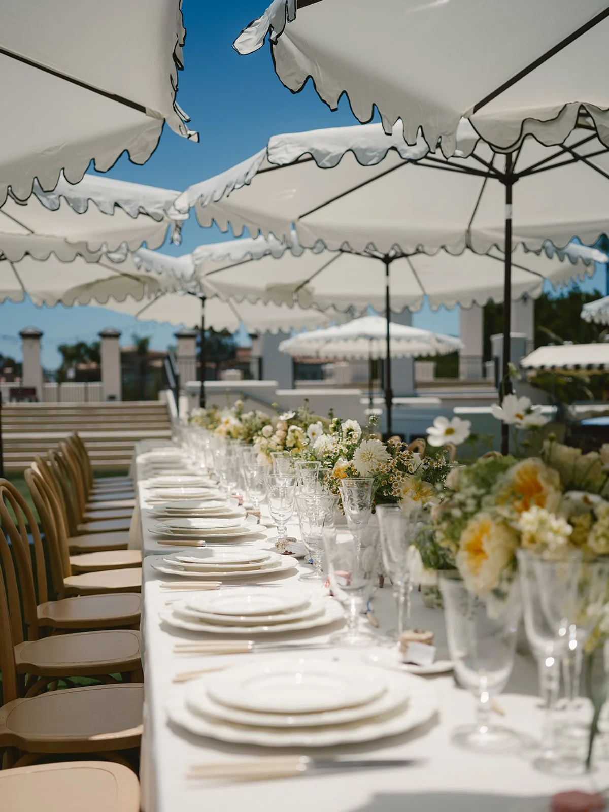Long outdoor dining table set with white plates, glassware, and floral centerpieces under white umbrellas on a sunny day.