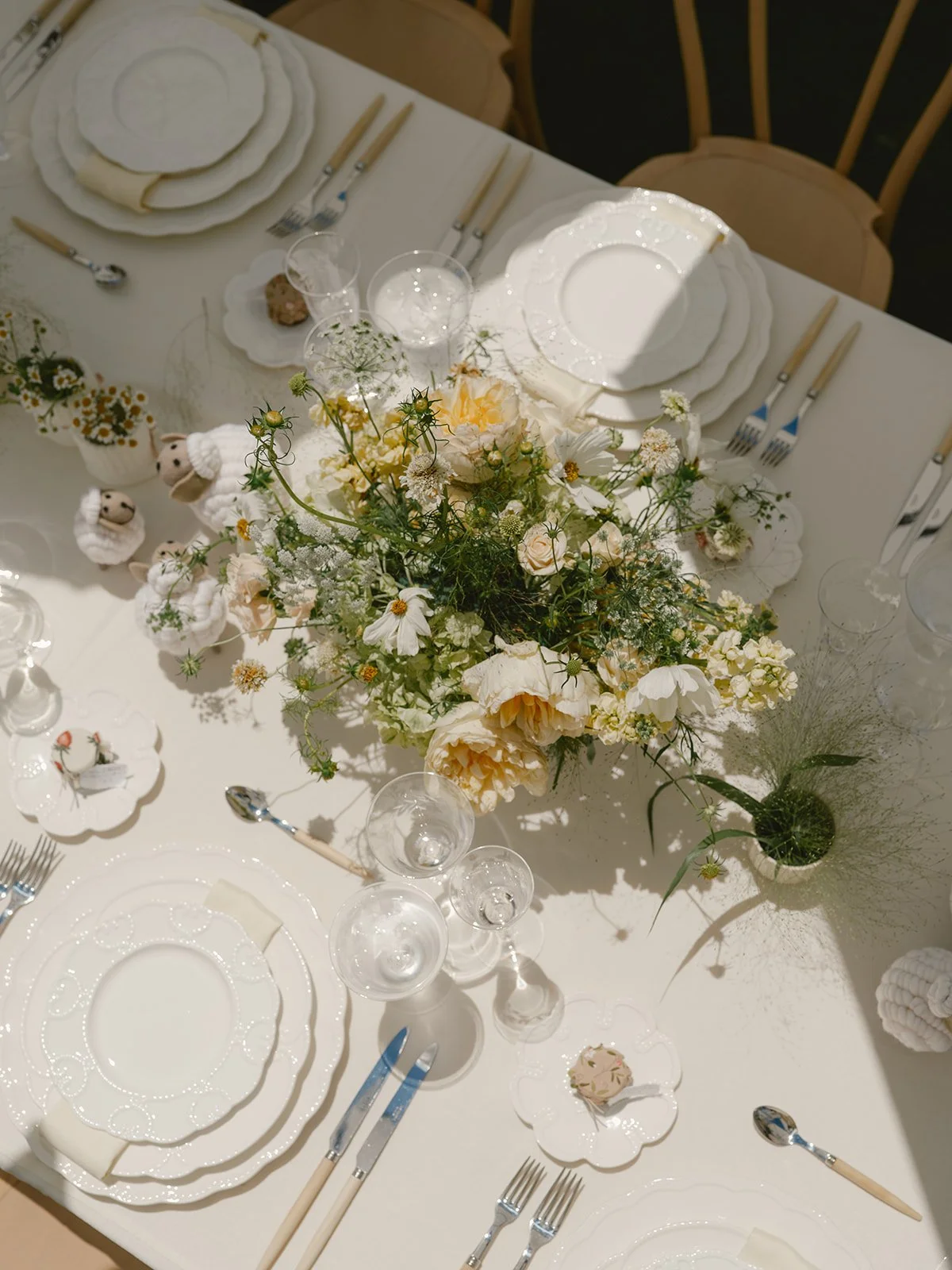 Table set for a formal occasion with white plates, silverware, and glassware, decorated with a central floral arrangement of white and cream-colored flowers and small plush lamb decorations.