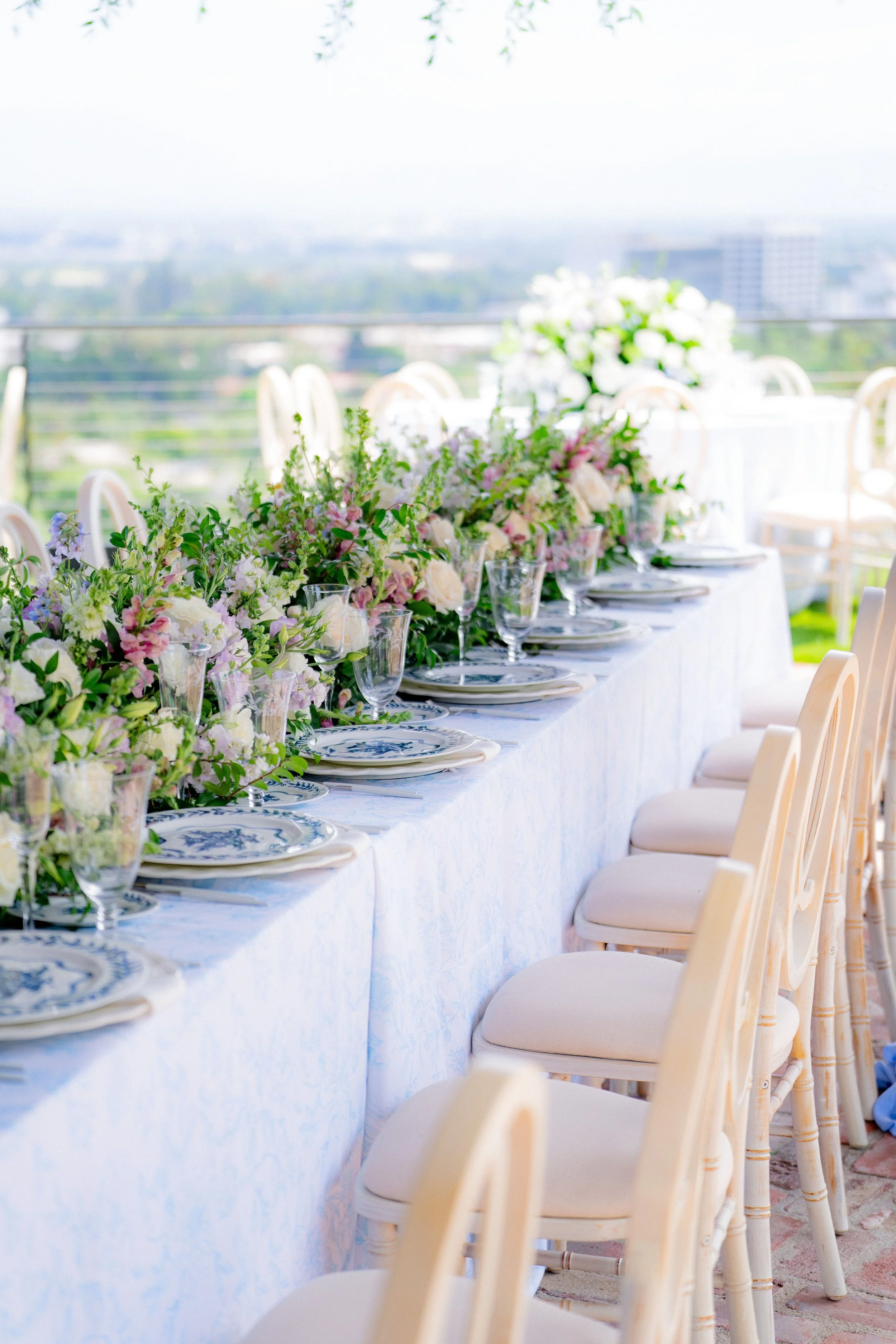 Elegant outdoor dining table set with floral centerpiece, plates, and glassware under a canopy with city view in the background.
