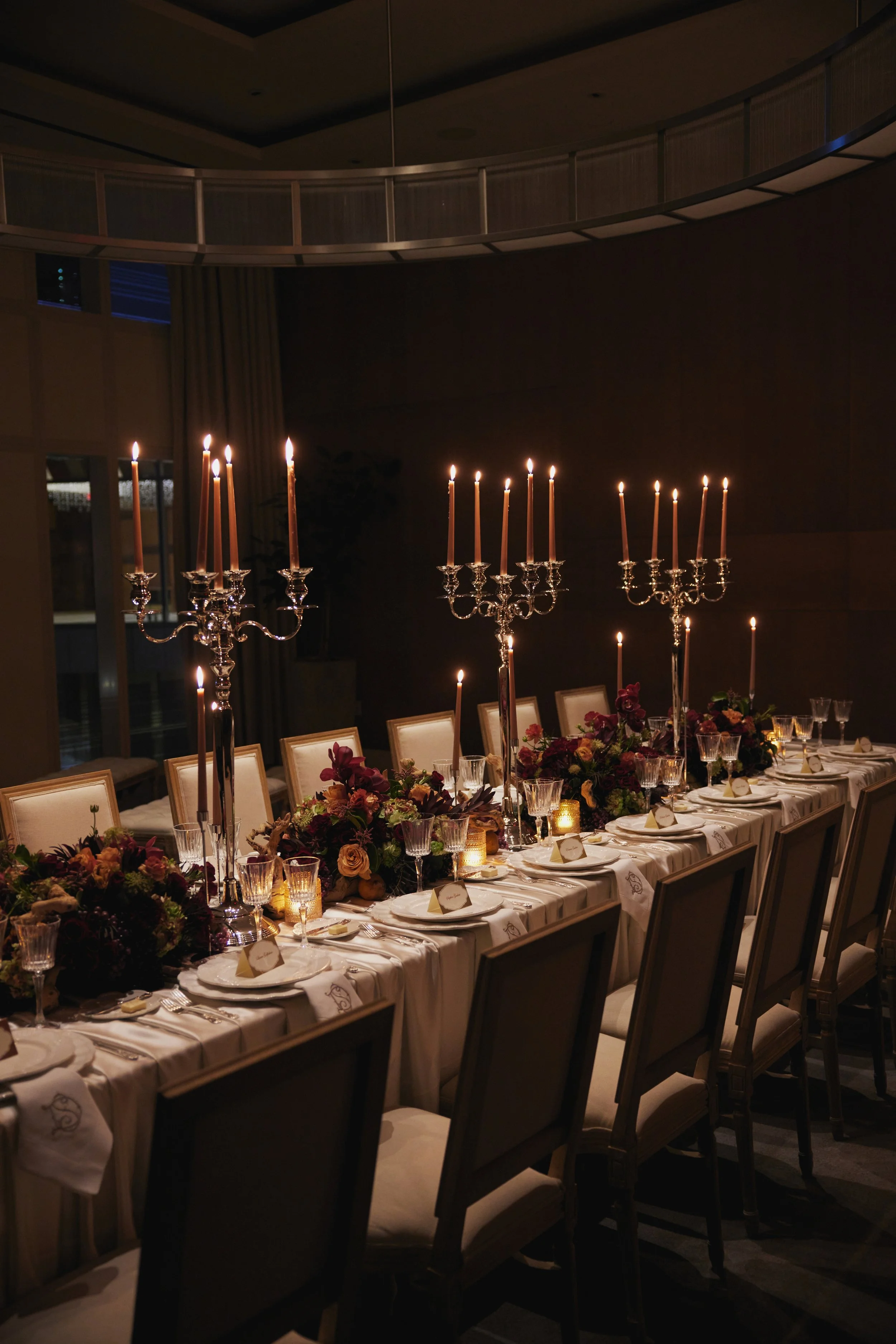 Elegant banquet table with white tablecloth, floral centerpieces, tall candleholders with lit candles, and place settings in a dimly lit room.