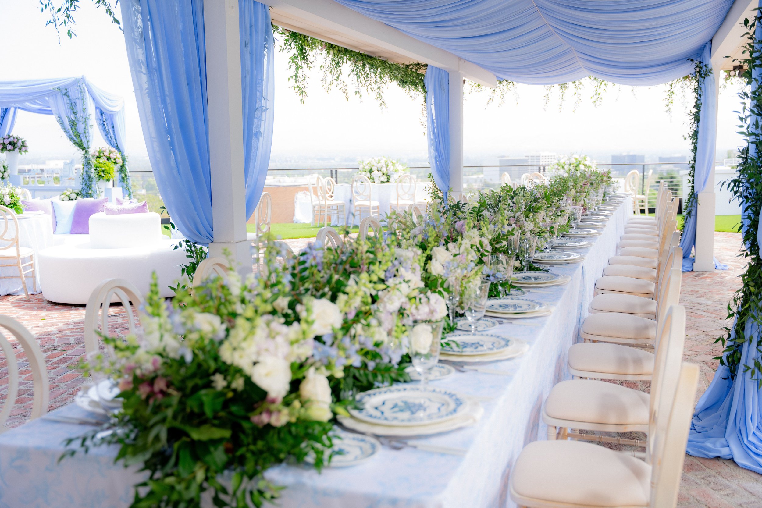 A beautifully decorated outdoor event space with a long banquet table adorned with floral centerpieces, set with plates and glasses, under a blue draped canopy and surrounded by white chairs, overlooking a cityscape.